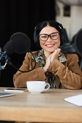happy asian radio host in glasses and headphones smiling near cup and smartphone on desk