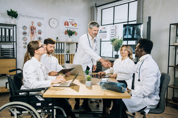 Fototapeta premium Bearded caucasian doctor shaking hands and greeting his multi ethnic colleagues during medical seminar at office. Female wheelchair user working with colleagues on common project.