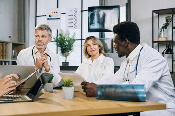 Obraz premium International group of five qualified doctors sitting together at table and using modern gadgets during discussion on patient's clinical case. Concept of people, health care and tecnology.