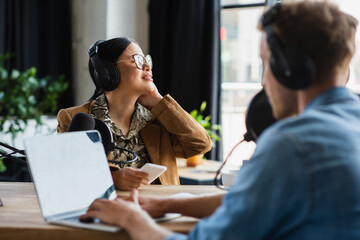 smiling asian radio host in headphones holding smartphone near blurred colleague typing on laptop