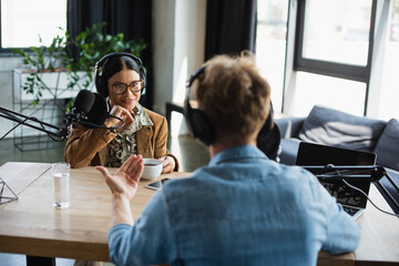 happy asian radio host in eyeglasses and headphones looking at blurred colleague during podcast