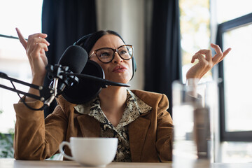 asian radio host in eyeglasses and headphones gesturing near cup of coffee on desk