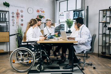 Aged male doctor with x ray scan in hands doing report to his international colleagues at office room. Multiracial practitioners cooperating for common medical research. One woman using wheelchair.