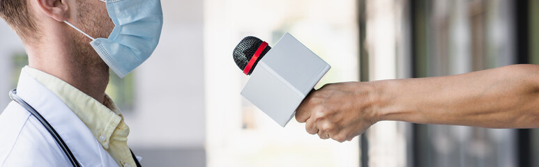 Obraz premium cropped view of reporter holding microphone near doctor in medical mask, banner