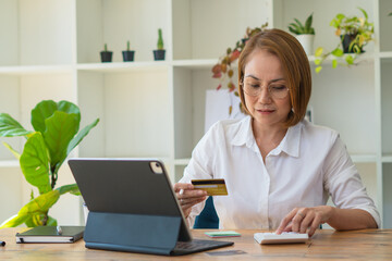 Serious focused middle aged female accountant in eyewear working from home providing outsource bookkeeping service, sitting at table with portable computer, calculating finances, doing paperwork