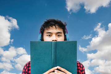 Asian young guys reading a book or listening an audiobook against a blue sky and clouds - Asian...