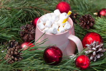 Close-up of a pink mug with hot beverage, marshmallow and straw in branches of a fir tree with red toy balls and cones. Christmas and New Year's holiday decorations