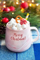 A mug with the text Merry Christmas with marshmallow, pretzel, caramel and straw on a plaid on a background of branch of a Christmas tree and bokeh lights. New Year holiday decorations. Vertical shot