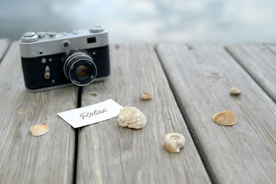 Wooden Background with a retro camera and seashells. A postcard with the words Relax lies on wooden planks on the beach. Empty old wooden table background with a lake in the background. Still life 