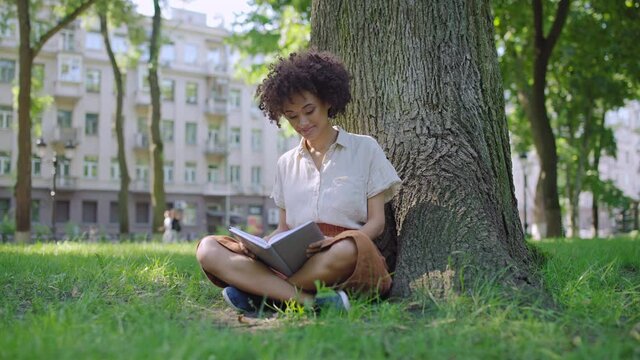 Beautiful young woman reading, sitting under tree, enjoying fresh air in park