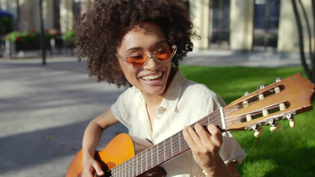 Cheerful young biracial woman with curly hair playing guitar, singing on street