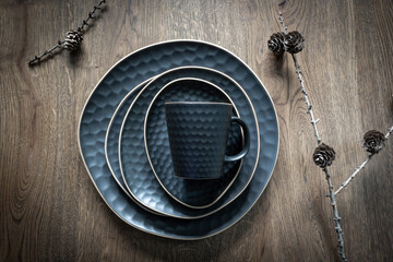 Dark blue set of dishes: a mug, saucer, plates, bowl and dry larch cones on twigs on wooden background. Christmas table decoration, magical holidays