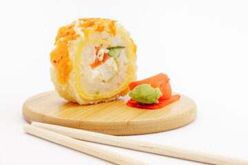 Still life with hot fried sushi roll on a round wooden plate with chopsticks, pickled pink ginger and green wasabi isolated on white background. Tempura roll with omelet, chicken and cucumber