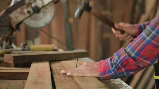Close up of carpenter man or joiner wears plaid shirt standing work nail hammer into wood plank for build furniture in workplace at factory. Concept of small business and owner startup about woodwork.