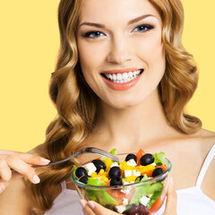 Square image of cheerful smiling woman holding salad with cheese and fresh vegetables, black olives, over yellow background. Blond girl at studio. Keto ketogenic diet, healthy eating. Greek salad.