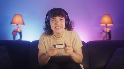 Portrait of excited young gamer woman sitting on couch, playing a video game with wireless controller on console at home. Young gamer girl wearing headphone © mahirkart