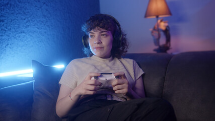 Young curly haired gamer girl with headphone sitting on sofa relaxing and playing a video game with wireless controller. Neon light background © mahirkart