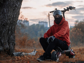 A young man rides an electric scooter in the fall wearing a helmet, balaclava and gloves. Autumn...