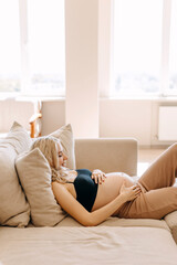 Fototapeta premium Pregnant woman wearing cozy home wear, lying on a sofa in a light interior, holding hands on belly.