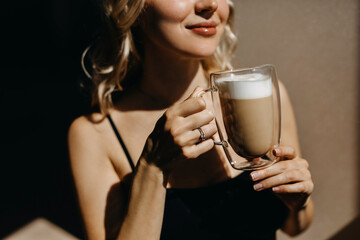 Closeup of a woman drinking coffee from a transparent double glass cup, in sunlight, smiling.