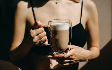 Closeup of a woman holding a transparent glass cup of coffee with latte in sunlight.