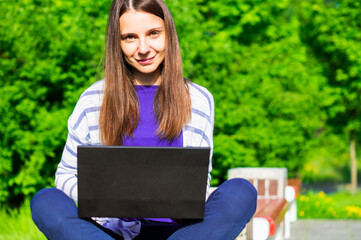 Beautiful young woman sitting in public park and working on laptop computer. Portrait of smiling woman studying on laptop at wile sitting on bench in park