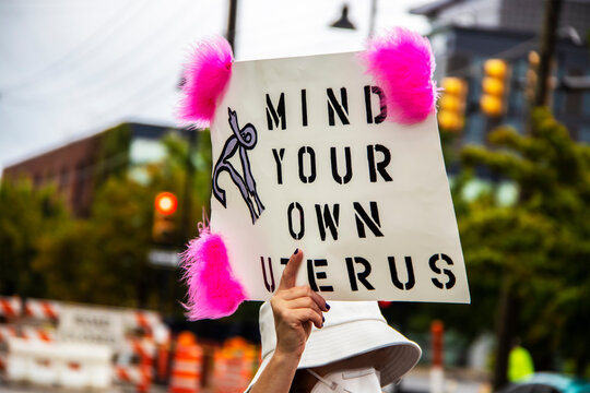 Woman in hat and mask at Reproductive Justice March holds up sign Mind Your Own Uterus