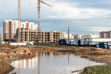 A large puddle after rain at the construction site of a large residential facility. Reflection in the water of the construction site and cranes against the background of the sunset sky.