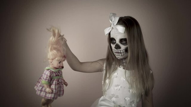 A little girl in a white dress with a painted face holds a bloody doll in her hand and looks at it.