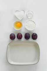 Step by step recipe for baking sweet plum pie. Ingredients set for cooking. Flour, eggs, sugar, plums for sweet pastries on white background. Top view, copy space. Culinary light background. 