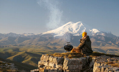 A man cooks on a compact charcoal grill on the Bermamyt plateau with a stunning view of Elbrus. Travel photo. - obrazy, fototapety, plakaty