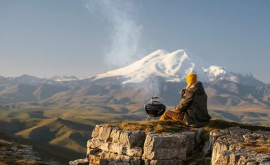 Fotobehang A man cooks on a compact charcoal grill on the Bermamyt plateau with a stunning view of Elbrus. Travel photo. © YULIYA