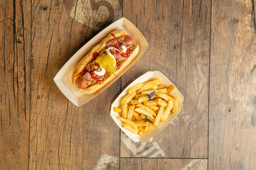 Top view image of hot dog with crispy onion, cooked sausage, fried bacon, mayonnaise, mustard and ketchup garnished with french fries in take away packages