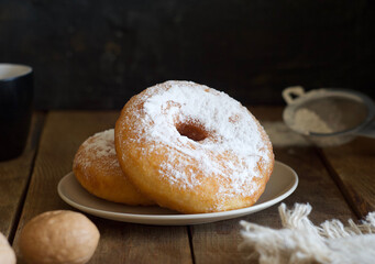 Two donuts with powder lie on a saucer on a wooden table. Nearby are a sieve, towel, walnut and mug. Rustic style
