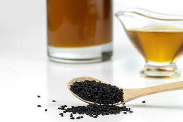 Black cumin seeds on a wooden spoon on a background of a glass gravy boat and of a bottle with black seed oil. Selective focus.