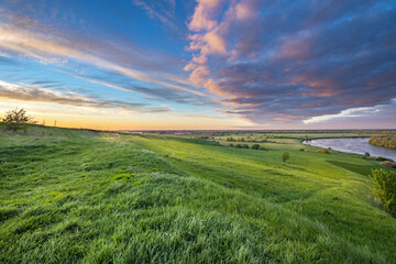 Scenic view of a beautiful summer river sunset with reflection in the water, calm water. Bright colorful cloudy sky. Lush green grass in the foreground.