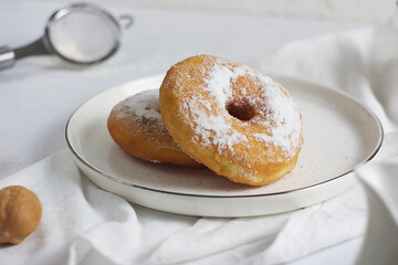 A pair of donuts in a plate on a gray table. Behind are a sieve, a walnut and a white towel