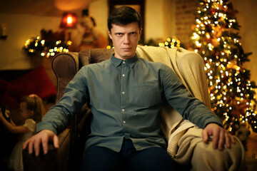 Obraz premium Portrait of a funny serious young father in an armchair, against the background of a christmas tree
