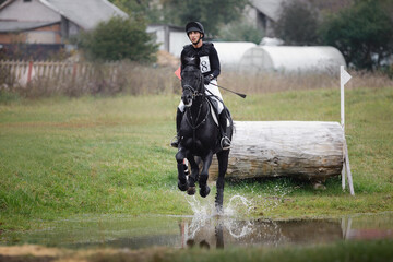 rider man galloping fast in water pond on black stallion horse during eventing cross country competition in autumn