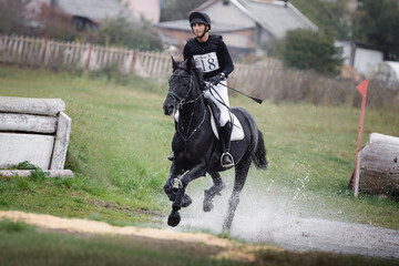 rider man galloping fast in water pond on black stallion horse during eventing cross country...