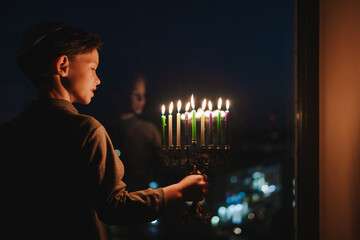 The child lights the menorah for Hanukkah on the windowsill. The boy in the kippah sitting by the...