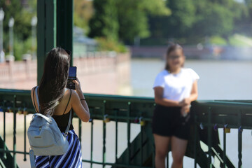 Dziewczyna fotografuje koleżankę telefonem komurkowym, smartfonem na moście. - obrazy, fototapety, plakaty
