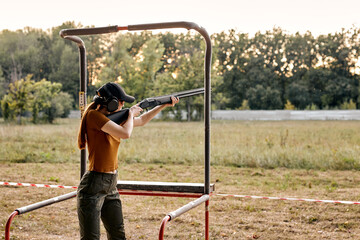 young caucasian woman in goggles and headset aiming rifle at side, ready to shoot in an outdoor range. Skilled experienced female side view. Firearms for sports shooting, hobby. - obrazy, fototapety, plakaty