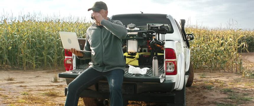 HANDHELD 50s Caucasian male farmer sitting on a back of a truck, checking his laptop while having a sandwich early in the morning