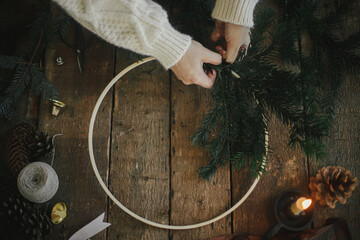 Fototapeta Hands making modern christmas wreath with fir branches on rustic table with round wooden hoop, scissors, thread, candle, pine cones. top view. Atmospheric moody image. Winter holidays preparation obraz