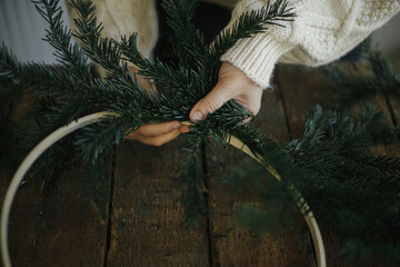 Hands making modern christmas wreath with fir branches on rustic table with round wooden hoop. Atmospheric moody image. Winter holidays preparation. Making minimalistic xmas wreath