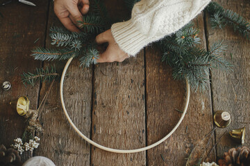 Hands in cozy sweater making modern christmas wreath with fir branches, herbs, bells, round wooden hoop on rustic table. Atmospheric moody image. Making xmas boho wreath - Powered by Adobe