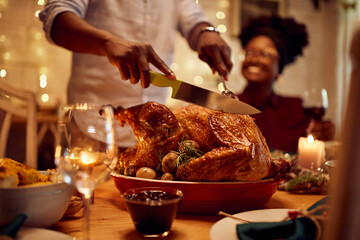 Close-up of African American man carves roasted Thanksgiving turkey at dining table. - obrazy, fototapety, plakaty