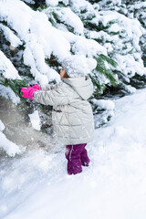 Fototapeta na wymiar child stands in a snowdrift near the Christmas trees and shakes the snow from the branches of trees. Winter games for children with snow. Happy child in a snowdrift