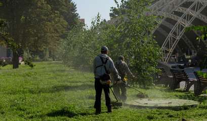 Two people with a manual lawn mower mows the grass. A man using a gasoline-powered string trimmer.